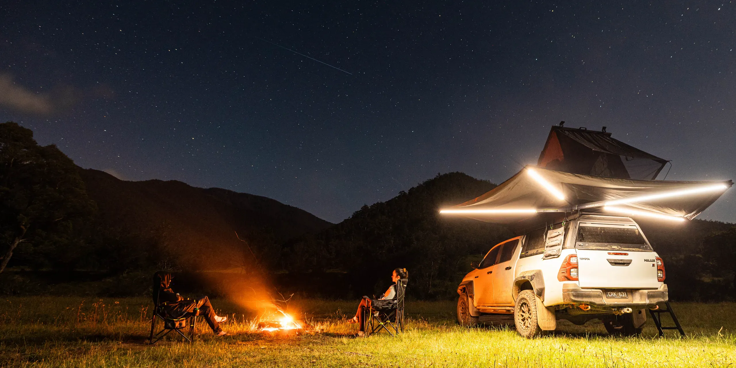 A rooftop tent and a lit awning set up beside a campfire under a night sky, with a fitted four wheel drive alongside
