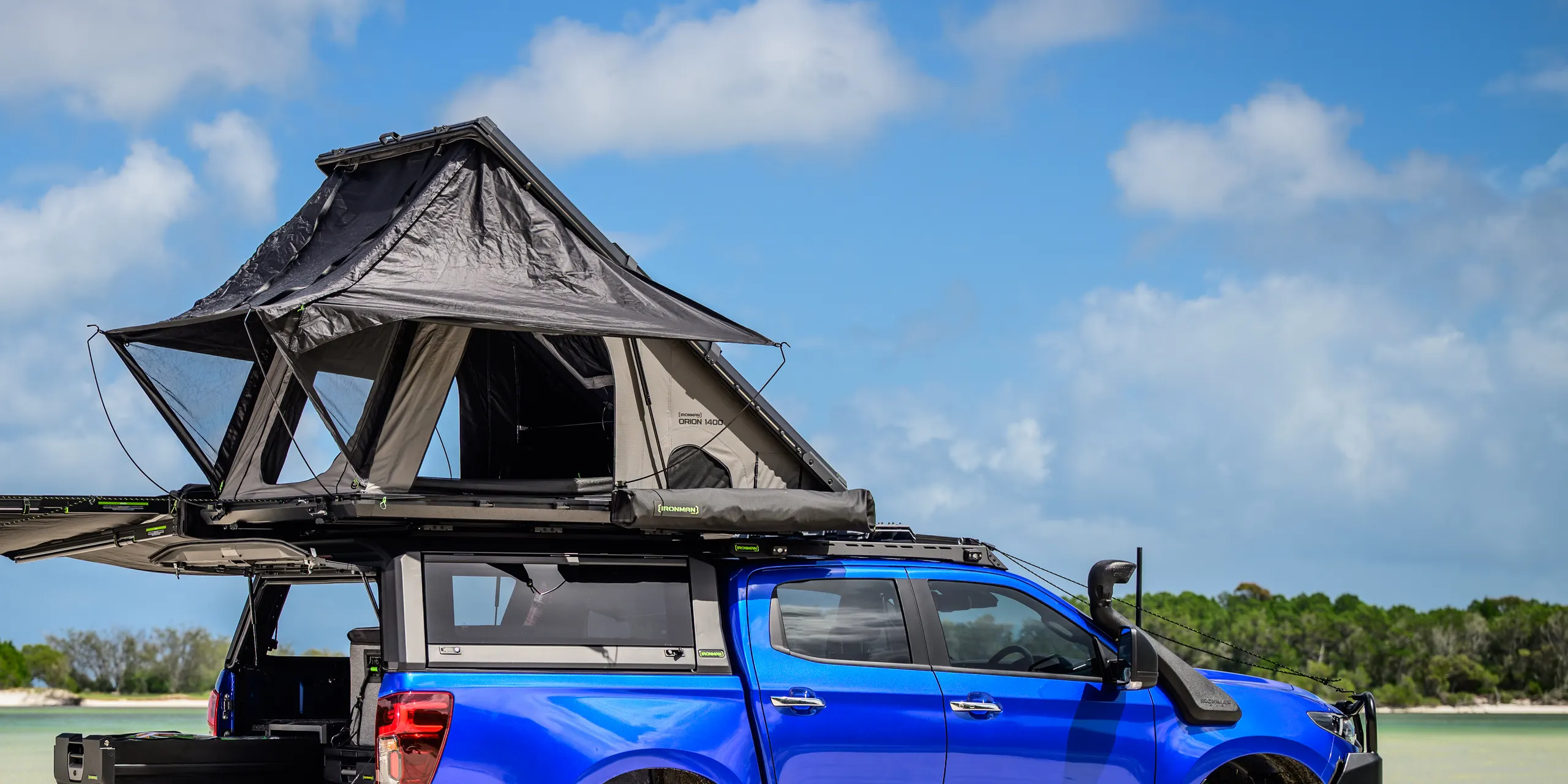 A rooftop tent set up on a roof platform above a ute canopy, with a folded awning alongside, on a beach