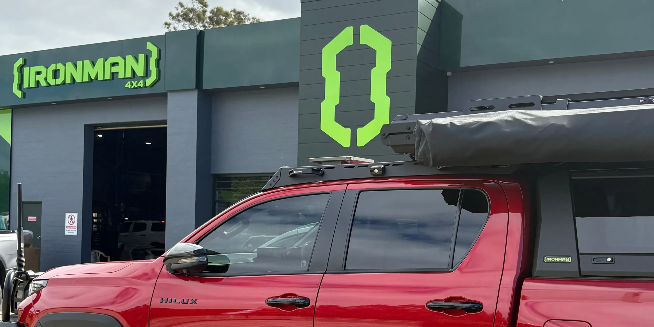 A red HiLux fitted with a roof platform, awning, canopy and side steps, outside the IRONMAN 4X4 store