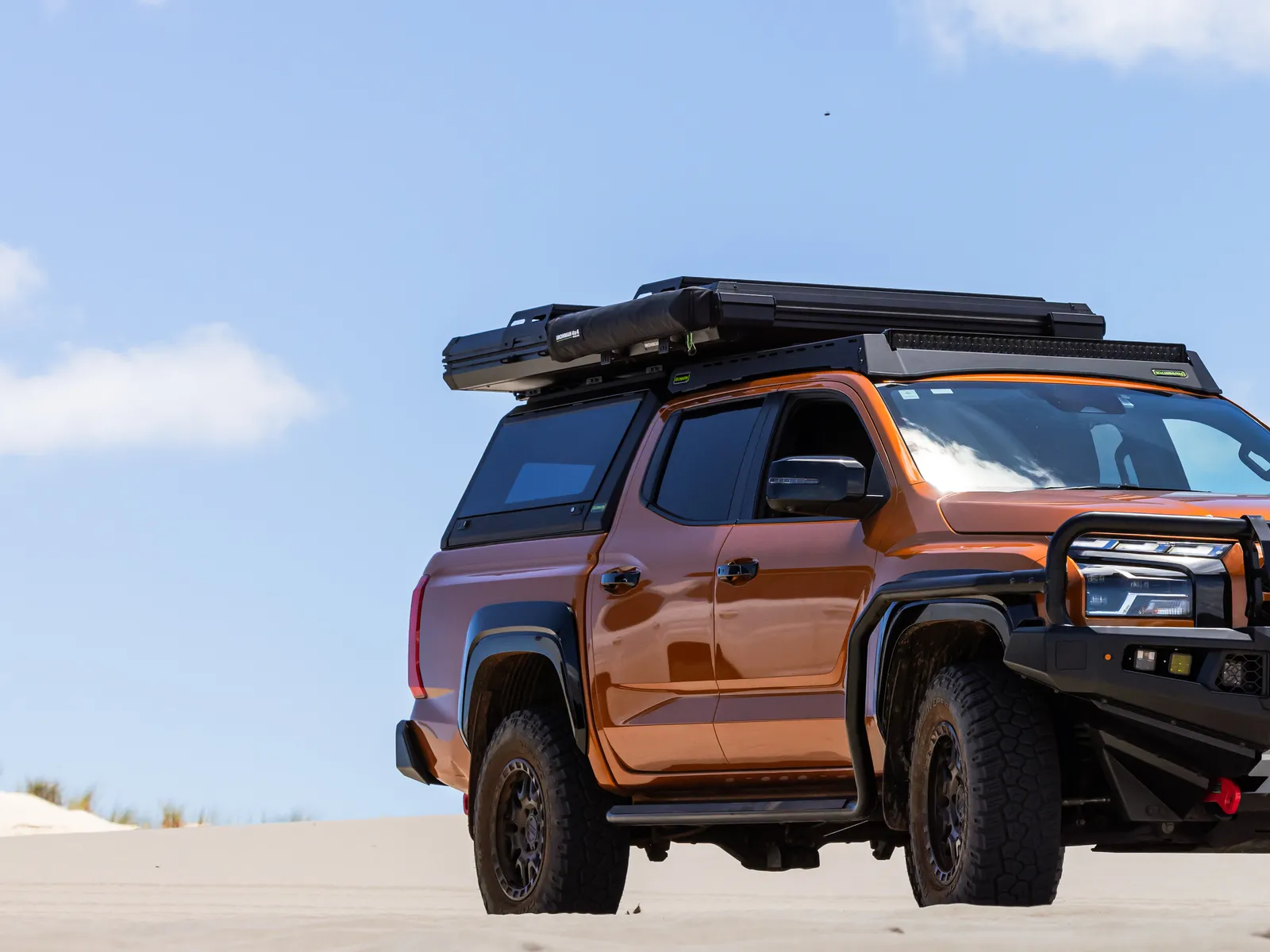 A four wheel drive fitted with a canopy, a roof platform and an awning, parked on a sand dune under a clear sky