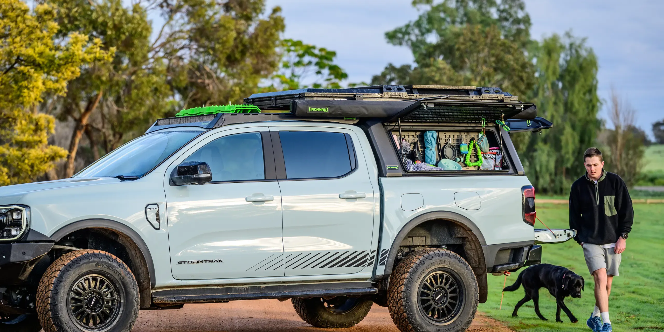 A dual cab ute fitted with a canopy, a roof platform, an awning and recovery boards, parked on a dirt track at dusk