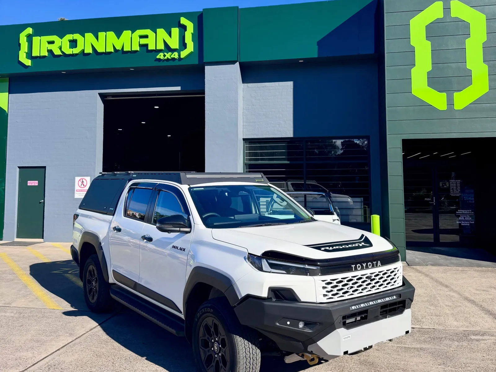 A white Toyota HiLux fitted with a canopy, roof platform and bar, parked outside the IRONMAN 4X4 store on Corrimal Street